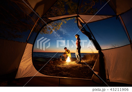 Young couple man and woman having rest at tourist tent and burning campfire on sea shore near forest Young couple man and woman having rest at tourist tent and burning campfire on sea shore near forest 43211906