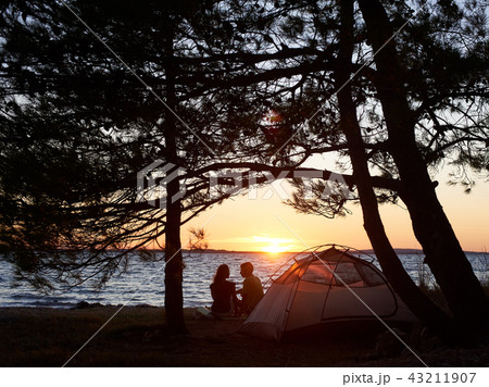 Young couple man and woman having rest at tourist tent and burning campfire on sea shore near forest Young couple man and woman having rest at tourist tent and burning campfire on sea shore near forest 43211907