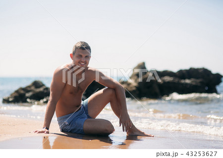 Handsome man in shorts sitting on the sand beach near rocks on the background of sea Handsome man in shorts sitting on the sand beach near rocks on the background of sea 43253627