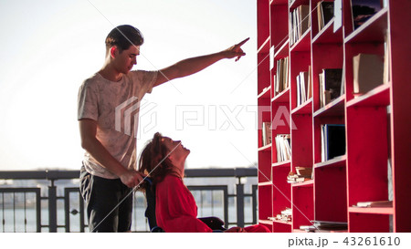 A young pretty girl is sitting in a wheelchair, her young man is standing next to her and pointing A young pretty girl is sitting in a wheelchair, her young man is standing next to her and pointing 43261610