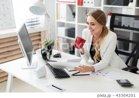 A young girl is sitting at the computer desk in the office and is holding a cup and documents A young girl is sitting at the computer desk in the office and is holding a cup and documents 43304201