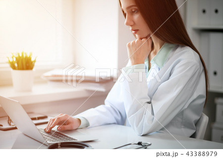 Young female doctor typing on laptop computer while sitting at the table near the window in hospital 43388979
