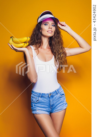 Woman in swimsuit and blue shorts holding banana and posing isolated over yellow background 43424902