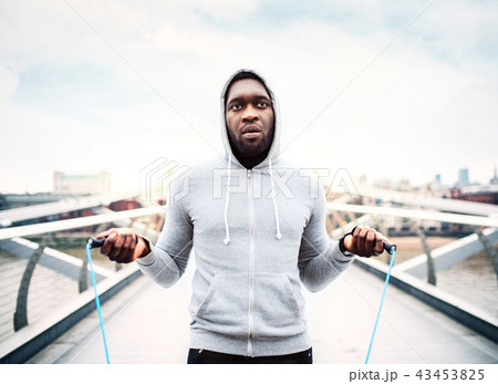 Young active black sportsman skipping with a rope in a city, wearing hoodie. 43453825