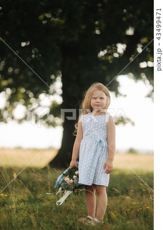 Little girl in sky blue dress with bouquet stand in field in front of big tree. Child smile and have 43499471