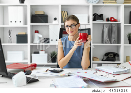 A young girl is sitting at a table in the office, holding a red cup in her hand and working with A young girl is sitting at a table in the office, holding a red cup in her hand and working with 43513018