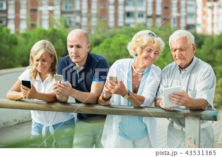 Positive mature couples holding gadgets in park Positive mature couples holding gadgets in park 43531365