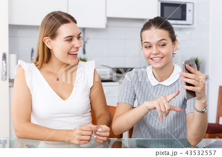 Positive young girlfriends looking at phone and chatting at table Positive young girlfriends looking at phone and chatting at table 43532557