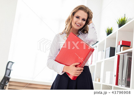 A beautiful young girl stands near a rack in the office and holds a folder with documents. A beautiful young girl stands near a rack in the office and holds a folder with documents. 43796908