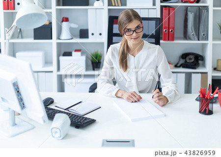 A young girl sits at a table in the office and holds a pencil in her hand. Before the girl lies an A young girl sits at a table in the office and holds a pencil in her hand. Before the girl lies an 43826959