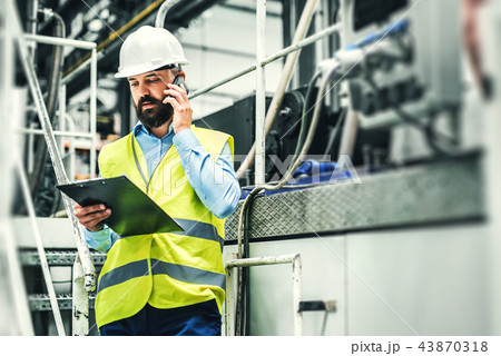 A portrait of an industrial man engineer with smartphone in a factory, working. A portrait of an industrial man engineer with smartphone in a factory, working. 43870318