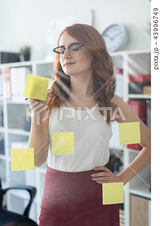 A beautiful young girl stands in the office near a glass on which stickers are pasted. The girl is A beautiful young girl stands in the office near a glass on which stickers are pasted. The girl is 43996749