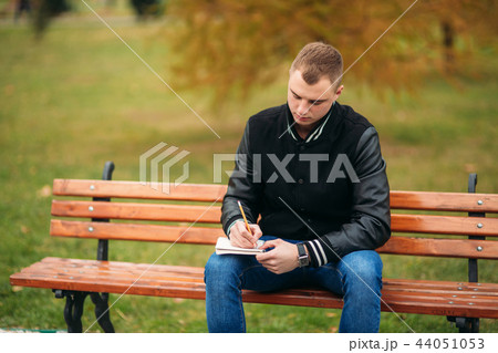 Student in a black jacket sits in a park on a bench writes down his thoughts in a notebook. Handsome 44051053