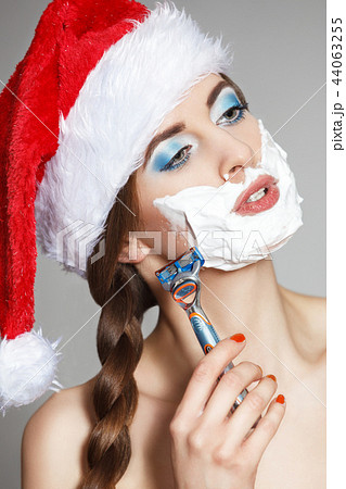 A beautiful woman in a Santa Claus hat shaves her face in a shaving foam with a man's razor. A beautiful woman in a Santa Claus hat shaves her face in a shaving foam with a man's razor. 44063255