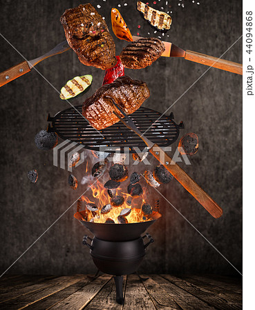 Kettle grill with hot briquettes, cast iron grate and tasty beef steaks flying in the air. 44094868