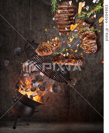 Kettle grill with hot briquettes, cast iron grate and tasty beef steaks flying in the air. 44094903