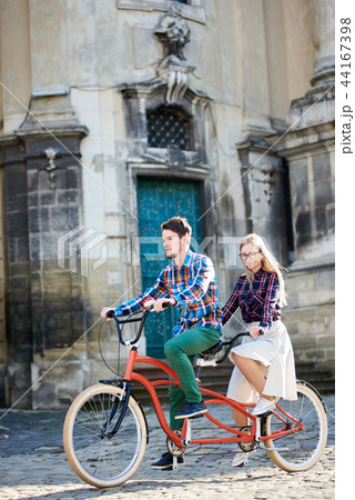 Young tourist couple, handsome man and pretty blond woman riding tandem bicycle along city street. 44167398