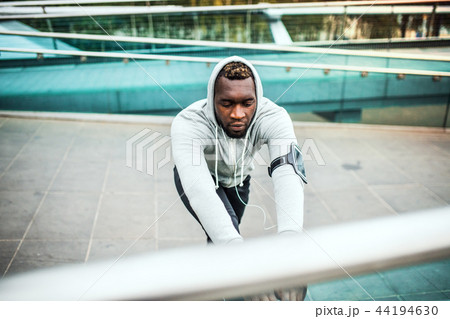Black man runner with smartphone in an armband on the bridge in a city, stretching. 44194630