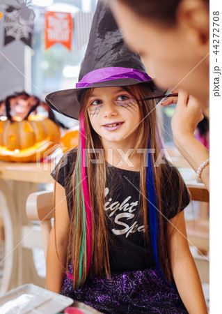 Dark-haired girl wearing wizard hat feeling happy getting Halloween makeup 44272708