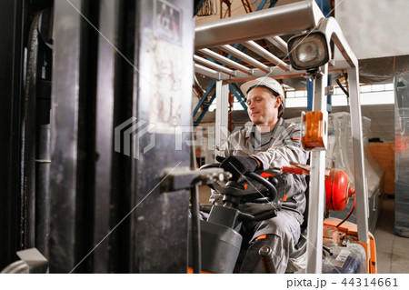 man driving a forklift through a warehouse in a factory. driver in uniform and protective helmet 44314661