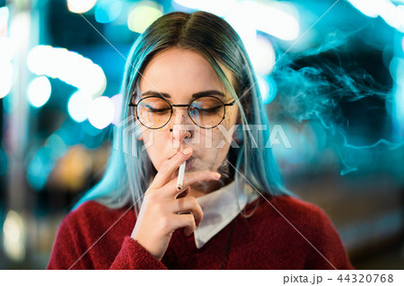 Millennial pretty girl with unusual dyed hairstyle smoking cigarette in amusement park at night 44320768