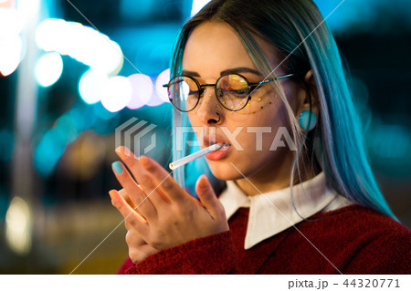 Millennial pretty girl with unusual dyed hairstyle smoking cigarette in amusement park at night 44320771