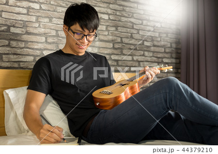 Happy young asian man playing ukulele sitting on bed in bedroom. Happy young asian man playing ukulele sitting on bed in bedroom. 44379210