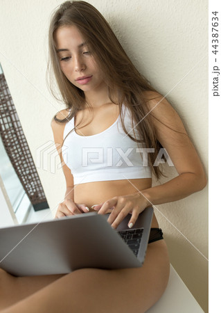 Young woman in a white top and panties sitting on the windowsill and using laptop. Young woman in a white top and panties sitting on the windowsill and using laptop. 44387634