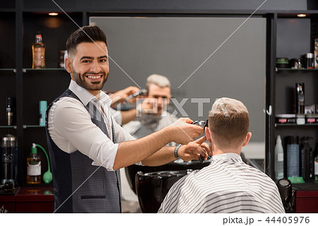 Smiling barber trimming client's stylish haircut and looking at camera. Smiling barber trimming client's stylish haircut and looking at camera. 44405976