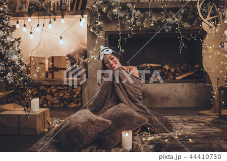 Beautiful asian woman is wearing sleeping mask and pajamas at home near christmas tree and fireplace Beautiful asian woman is wearing sleeping mask and pajamas at home near christmas tree and fireplace 44410730