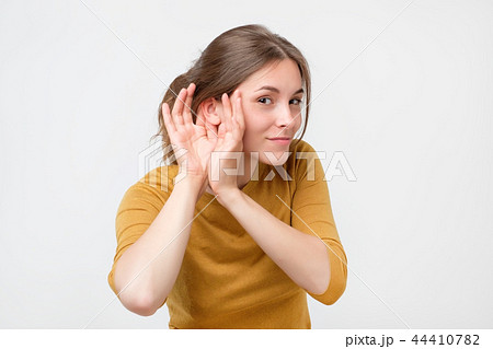 Young woman in yellow sweater listening to gossip about other with hand near ear. 44410782
