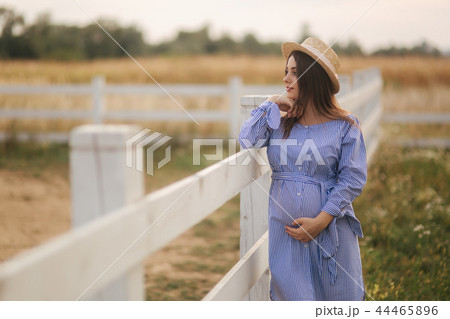 Beautiful pregnant woman in blue dress and knitted hat stand near the fance in the farm. She put 44465896