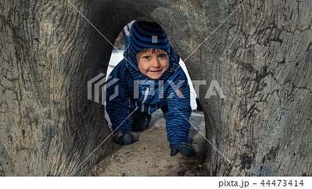 Beautiful kid in warm clothes and hat playing and crawls under the tree Beautiful kid in warm clothes and hat playing and crawls under the tree 44473414