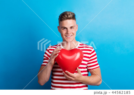A young man in a studio, holding red heart balloon in front of him. 44562185