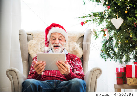 A portrait of senior man with tablet sitting on armchair at home at Christmas time. A portrait of senior man with tablet sitting on armchair at home at Christmas time. 44563298
