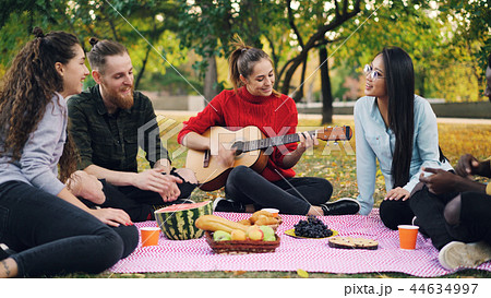 Charming young woman is playing the guitar sitting on blanket with friends on picnic, girls and guys 44634997