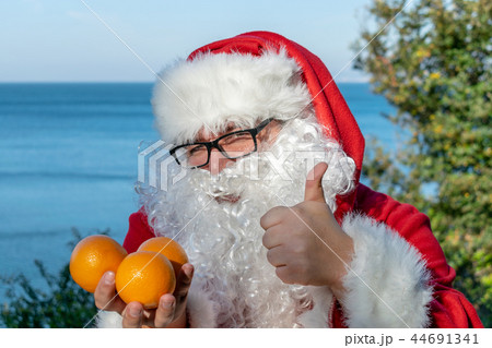 Fat man in glasses dressed as Santa holds tangerines on the ocean. Vacation and healthy lifestyle Fat man in glasses dressed as Santa holds tangerines on the ocean. Vacation and healthy lifestyle 44691341