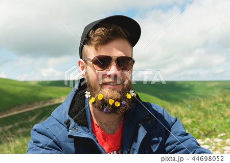 Close-up portrait of a cheerful bearded man in sunglasses and a gray cap with wildflowers in a beard Close-up portrait of a cheerful bearded man in sunglasses and a gray cap with wildflowers in a beard 44698052