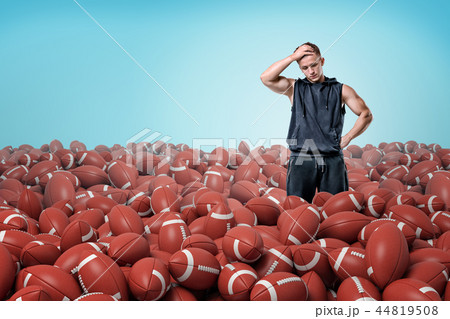 A thoughtful muscular man stands in a field of identical American football balls. A thoughtful muscular man stands in a field of identical American football balls. 44819508