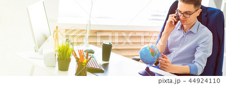 A young man sits in the office at a computer desk, talking on the phone and looking at the globe. 44924110