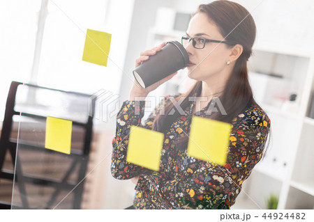 A young girl stands in the office near a transparent Board with stickers and drinks coffee. A young girl stands in the office near a transparent Board with stickers and drinks coffee. 44924482