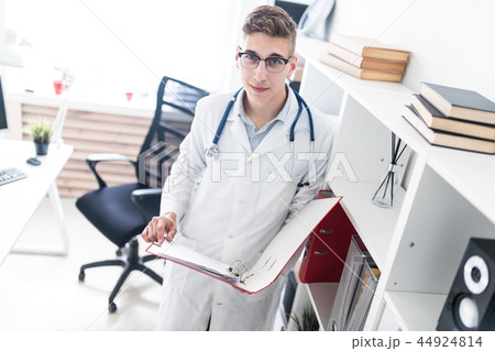 A young doctor in a white robe standing near the rack and holding a folder with documents. A young doctor in a white robe standing near the rack and holding a folder with documents. 44924814