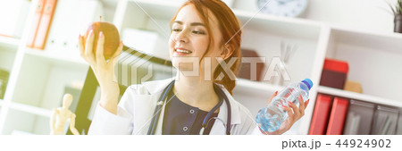 A beautiful young girl in a white robe sits at a table in the office and holds an apple and a bottle A beautiful young girl in a white robe sits at a table in the office and holds an apple and a bottle 44924902