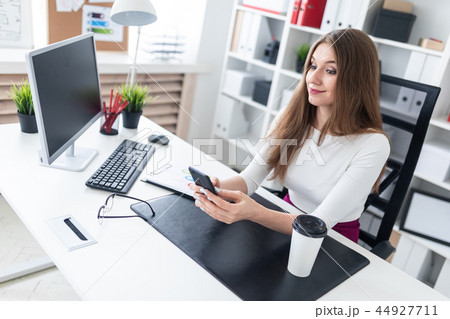 A young girl sitting at a table in the office and holding a phone. Next to the table is a white 44927711