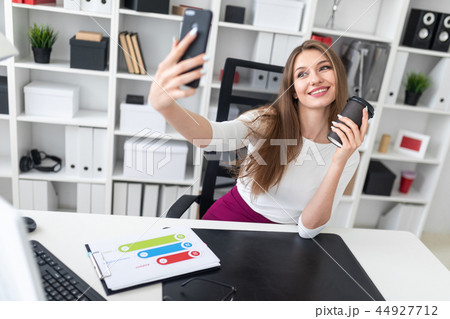 A young girl sitting at a table in the office and holding a glass of coffee and a phone. 44927712