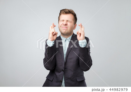 Handsome young man in suit looking at camera and keeping fingers crossed wishing some luck Handsome young man in suit looking at camera and keeping fingers crossed wishing some luck 44982979