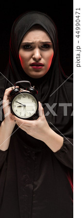 Portrait of beautiful serious scared frightened young muslim woman wearing black hijab holding alarm Portrait of beautiful serious scared frightened young muslim woman wearing black hijab holding alarm 44990471