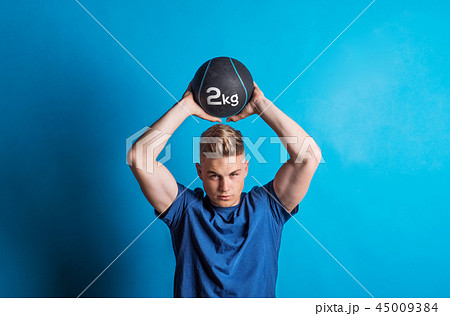 Portrait of a young man holding a heavy ball above his head in a studio. 45009384
