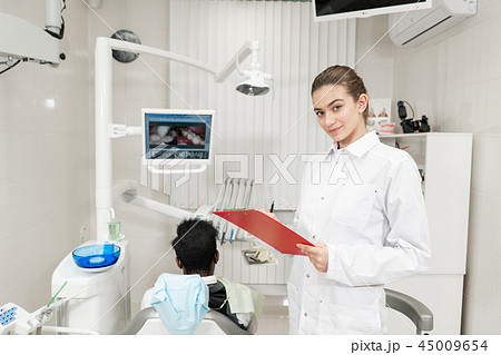 Charming beautiful girl dentist looks into the camera. Young African American male patient at chair 45009654