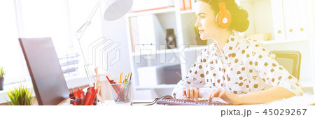 Beautiful young girl sitting in headphones at desk in office and typing on keyboard. 45029267